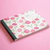 A floral-patterned notebook with a white cover, pink and red flowers, and green leaves, resting on a solid pink background. The cover features Japanese text and a dark green spine.
