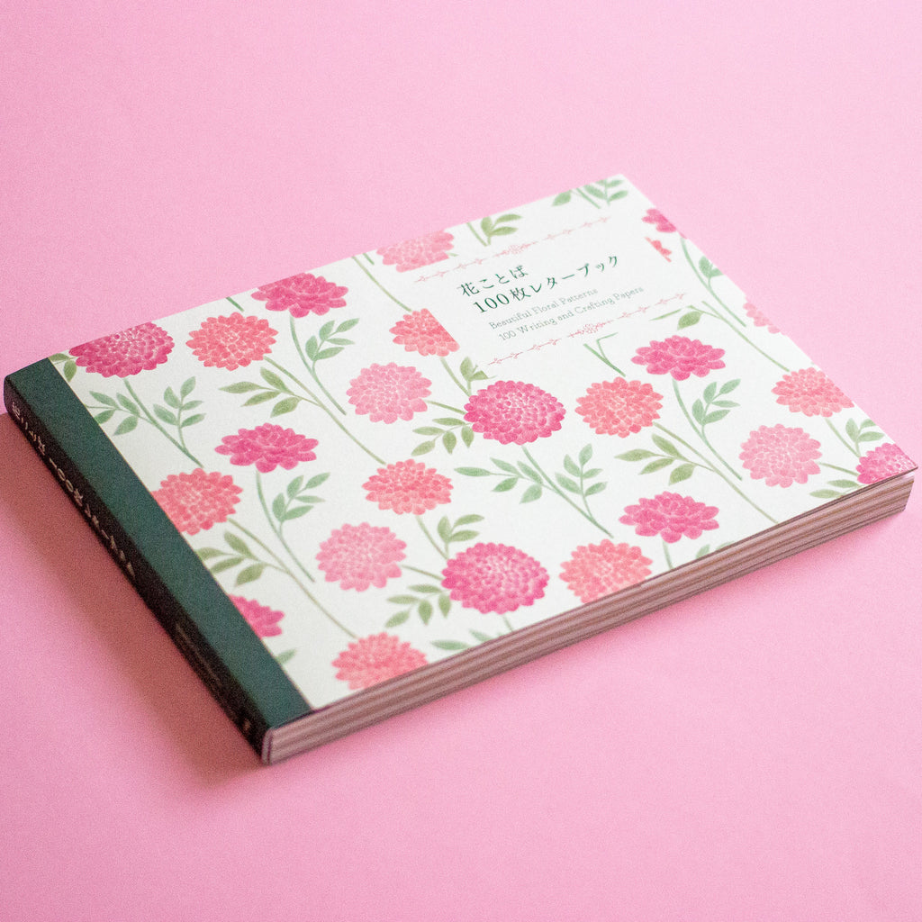 A floral-patterned notebook with a white cover, pink and red flowers, and green leaves, resting on a solid pink background. The cover features Japanese text and a dark green spine.