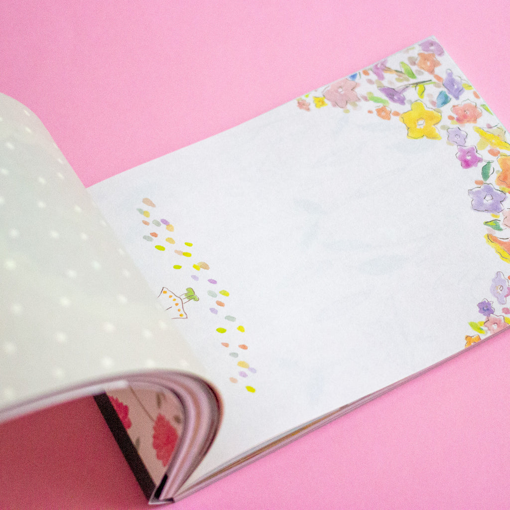 An open notebook on a solid pink background, showing a white page with a colourful floral border and small pastel polka dots.