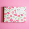 A floral notebook or art book with a white cover decorated with pink and red flowers and green stems, photographed on a solid pink background. The cover includes Japanese and English text.