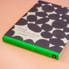 A book titled '100 Writing & Crafting Papers' by Kamawanu, featuring a black and white patterned cover with a bright green spine, photographed on a solid peach-pink background.