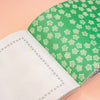 An open notebook with blank white pages on one side and a green cover on the other, decorated with a repeating white geometric star pattern, set against a solid peach-pink background.
