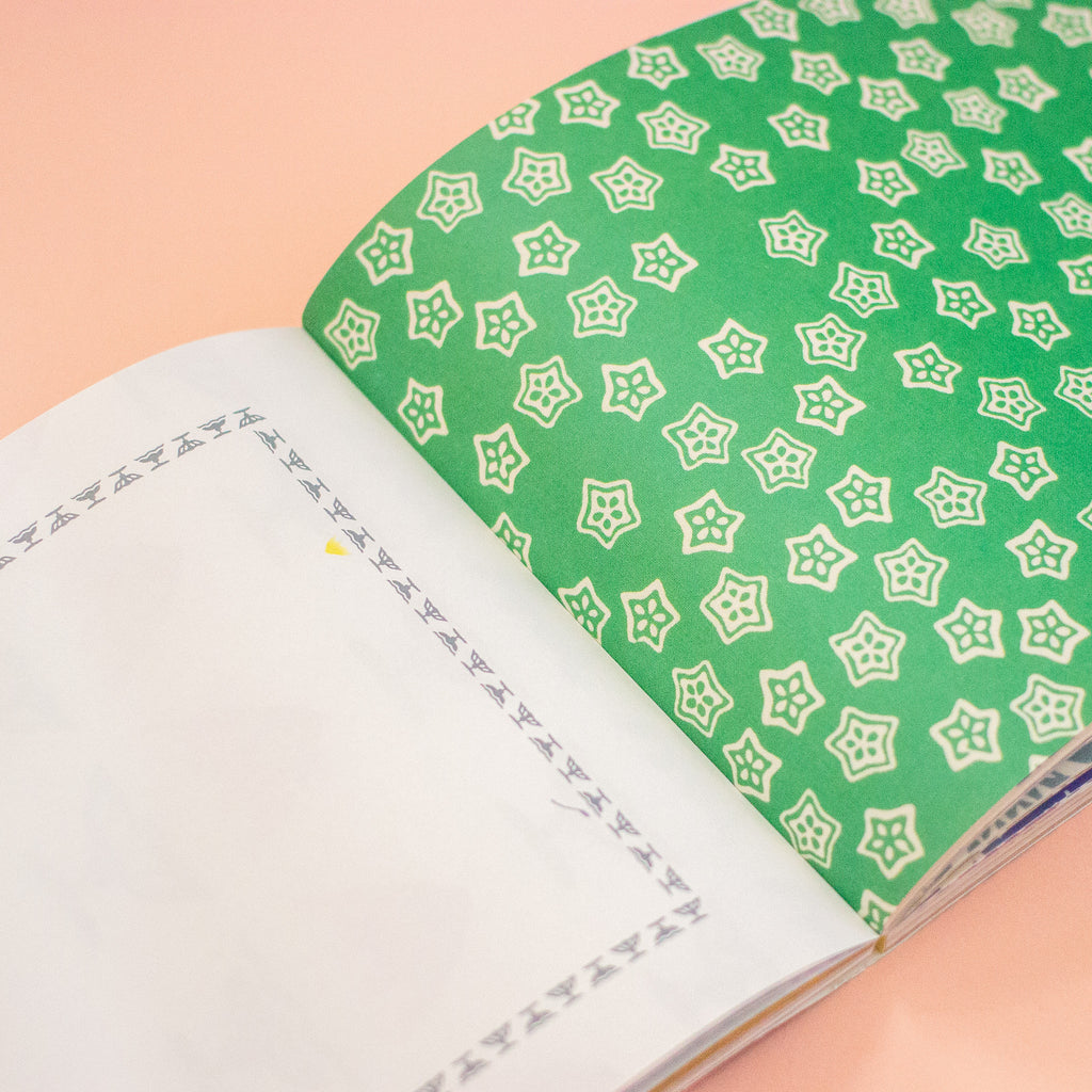 An open notebook with blank white pages on one side and a green cover on the other, decorated with a repeating white geometric star pattern, set against a solid peach-pink background.