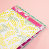 A stack of patterned paper sheets or cards on a soft peachbackground. The top sheet is white with yellow dotted floral clusters and green leaf motifs, and pink patterned sheets are visible underneath.