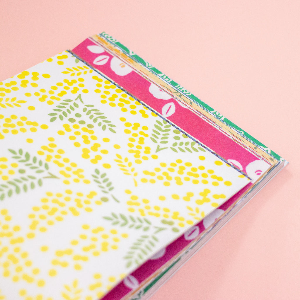 A stack of patterned paper sheets or cards on a soft peachbackground. The top sheet is white with yellow dotted floral clusters and green leaf motifs, and pink patterned sheets are visible underneath.