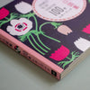A close-up angled shot of a Japanese book cover featuring a dark purple background with stylised white, pink, and green flowers, plus a pink spine band with Japanese text.