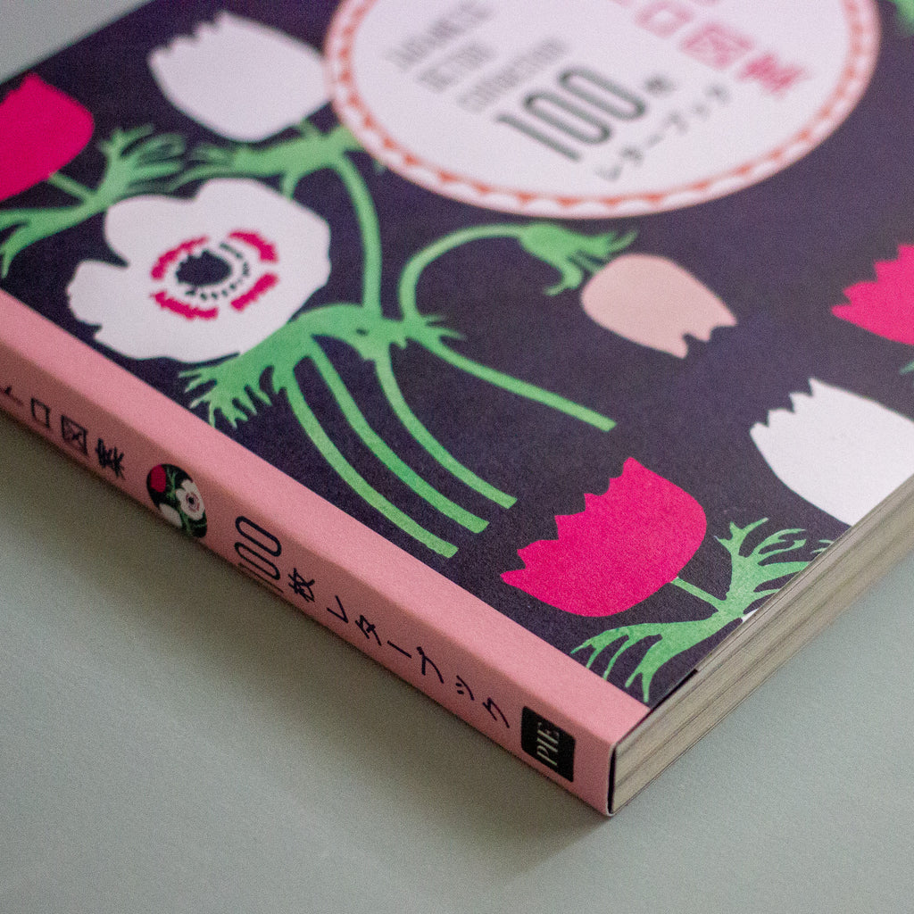 A close-up angled shot of a Japanese book cover featuring a dark purple background with stylised white, pink, and green flowers, plus a pink spine band with Japanese text.