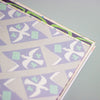 A close-up of a patterned paper product corner, showing a geometric design with white doves, green clovers, and light green envelopes on a lavender and grey background.