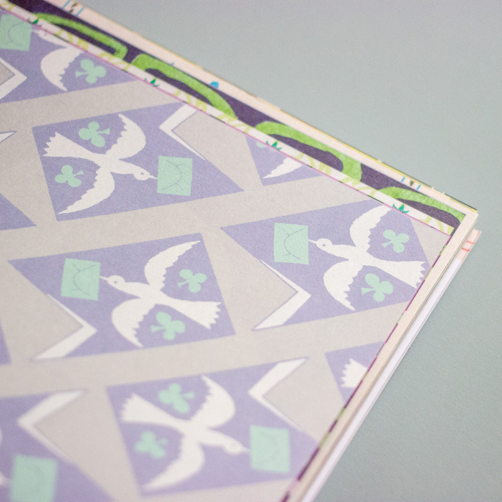 A close-up of a patterned paper product corner, showing a geometric design with white doves, green clovers, and light green envelopes on a lavender and grey background.