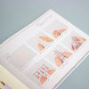 An open booklet or instruction manual showing printed steps for wrapping a phone with decorative paper, photographed on a pale blue background.