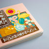 A close-up of a decorative book or notebook cover with a pastel pink border and an illustrated central panel featuring a yellow swan, turquoise gem, cream flowers, and ornate brown and blue motifs, photographed on a light grey background.