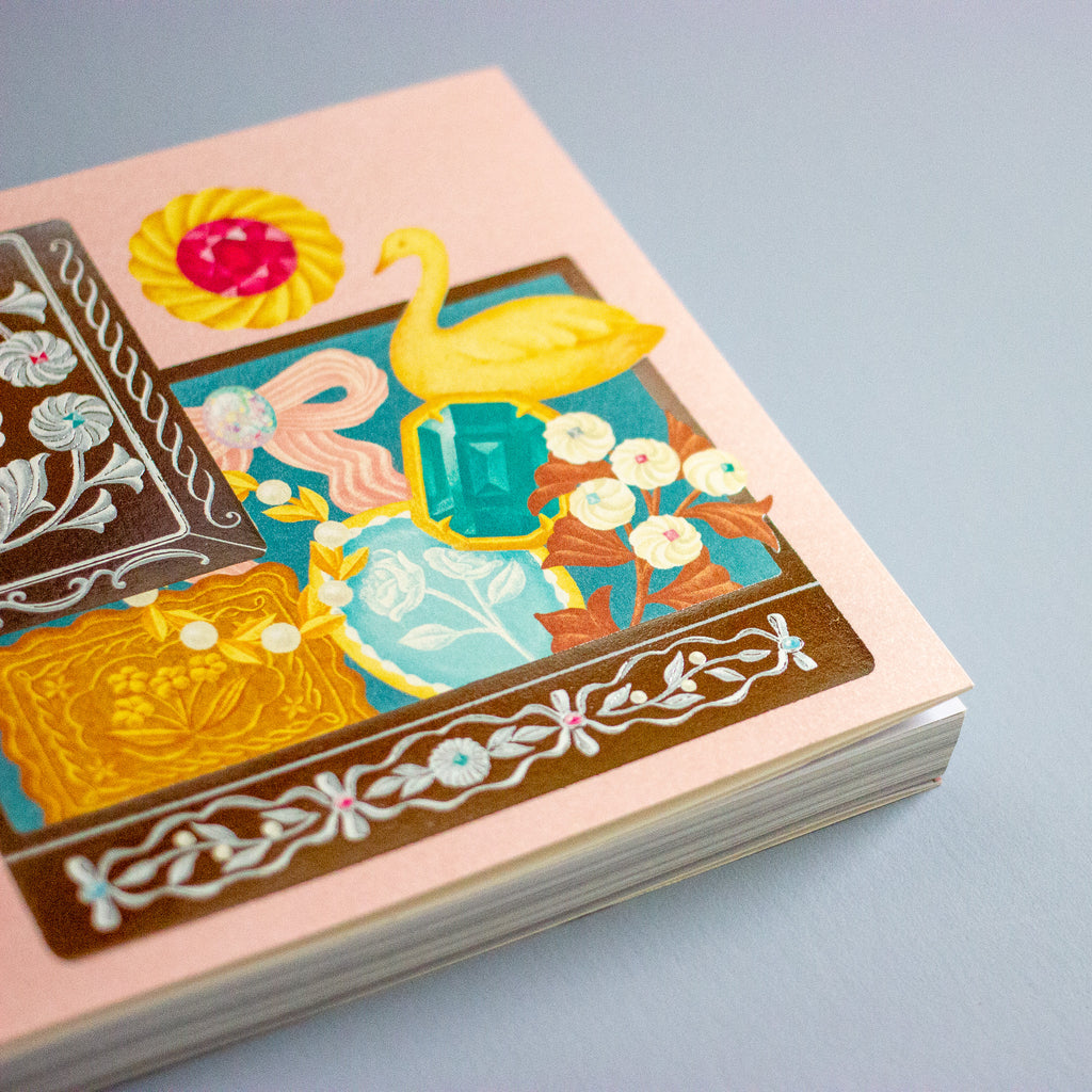 A close-up of a decorative book or notebook cover with a pastel pink border and an illustrated central panel featuring a yellow swan, turquoise gem, cream flowers, and ornate brown and blue motifs, photographed on a light grey background.