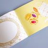 A close-up of a booklet page with a pale yellow background, gold decorative grid lines, and illustrations of pastries and a teapot, alongside a white ornate frame on the left, photographed on a blue-grey surface.