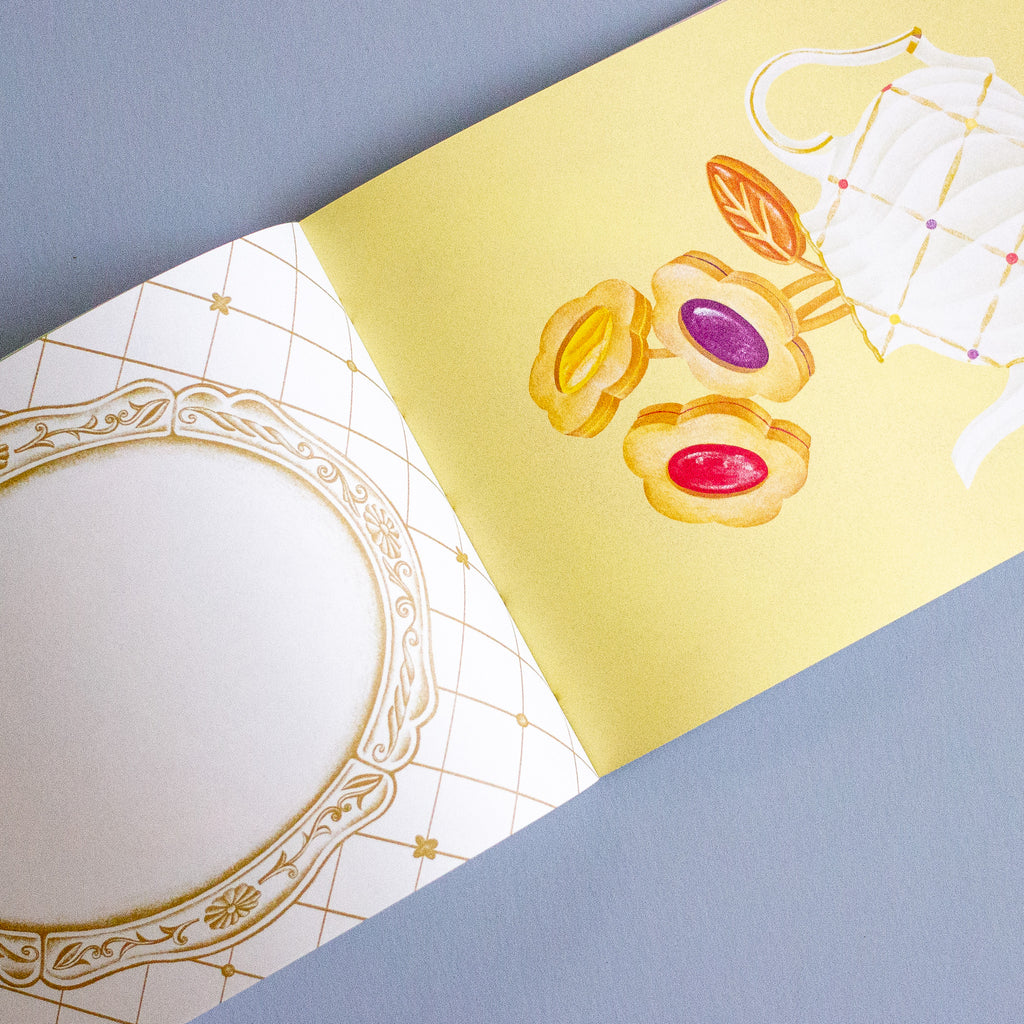 A close-up of a booklet page with a pale yellow background, gold decorative grid lines, and illustrations of pastries and a teapot, alongside a white ornate frame on the left, photographed on a blue-grey surface.