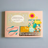 A pastel pink notebook or journal cover with decorative illustrated elements, including a swan, ribbon, flowers, and a gold ornamental band, photographed against a plain light blue background.