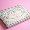 A decorative book of 100 patterned papers with classical floral designs, photographed at an angle on a solid pink background.