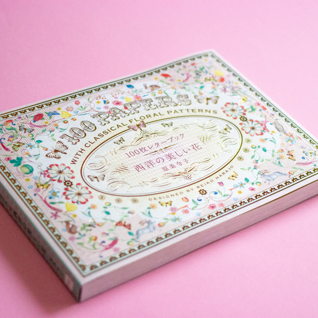 A decorative book of 100 patterned papers with classical floral designs, photographed at an angle on a solid pink background.