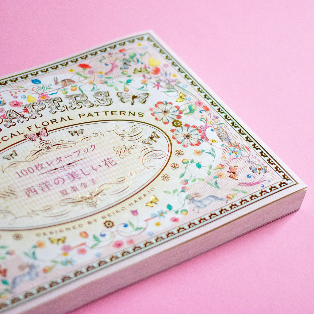 A decorative book cover with floral and butterfly illustrations, photographed on a solid pink background.