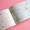 An open book on a solid pink background, showing a floral patterned cover on the right page and small scattered floral motifs on the left page.