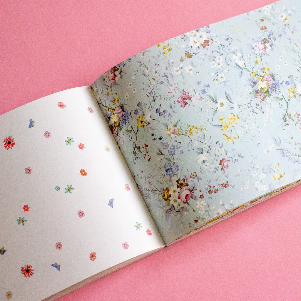 An open book on a solid pink background, showing a floral patterned cover on the right page and small scattered floral motifs on the left page.