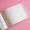 An open notebook on a solid pink background, showing lined writing pages with gold decorative borders and a floral page on the left.