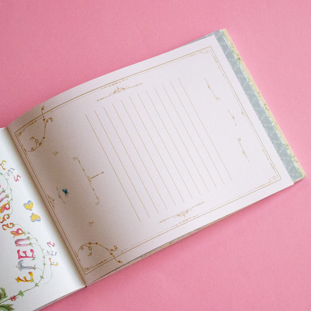 An open notebook on a solid pink background, showing lined writing pages with gold decorative borders and a floral page on the left.
