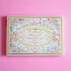 A decorative book cover with an ornate floral border and gold lettering reading '100 PAPER SHEETS' and 'WITH CLASSICAL FLORAL PATTERNS,' photographed on a solid pink background.
