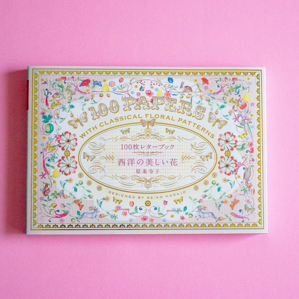 A decorative book cover with an ornate floral border and gold lettering reading '100 PAPER SHEETS' and 'WITH CLASSICAL FLORAL PATTERNS,' photographed on a solid pink background.