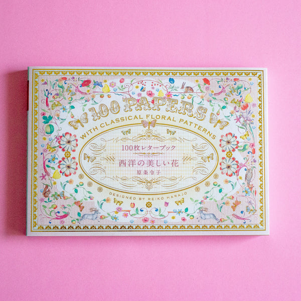 A decorative book cover with an ornate floral border and gold lettering reading '100 PAPER SHEETS' and 'WITH CLASSICAL FLORAL PATTERNS,' photographed on a solid pink background.