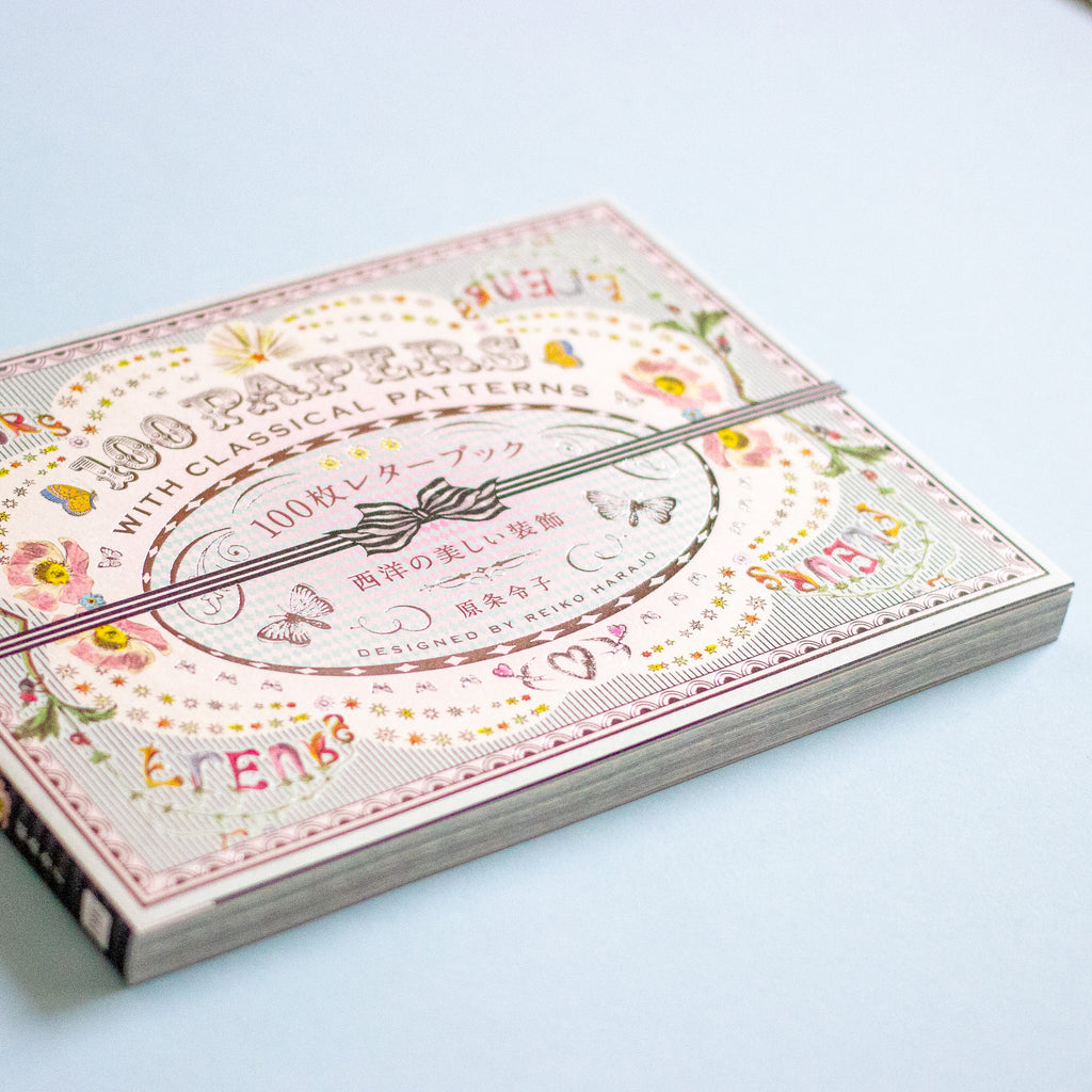 A book of vintage-style patterned papers in a decorative cover, photographed on a clean light blue background. The packaging features pink, beige, and gold floral and ornamental designs with text including '100 PAPERS WITH CLASSICAL PATTERNS'.