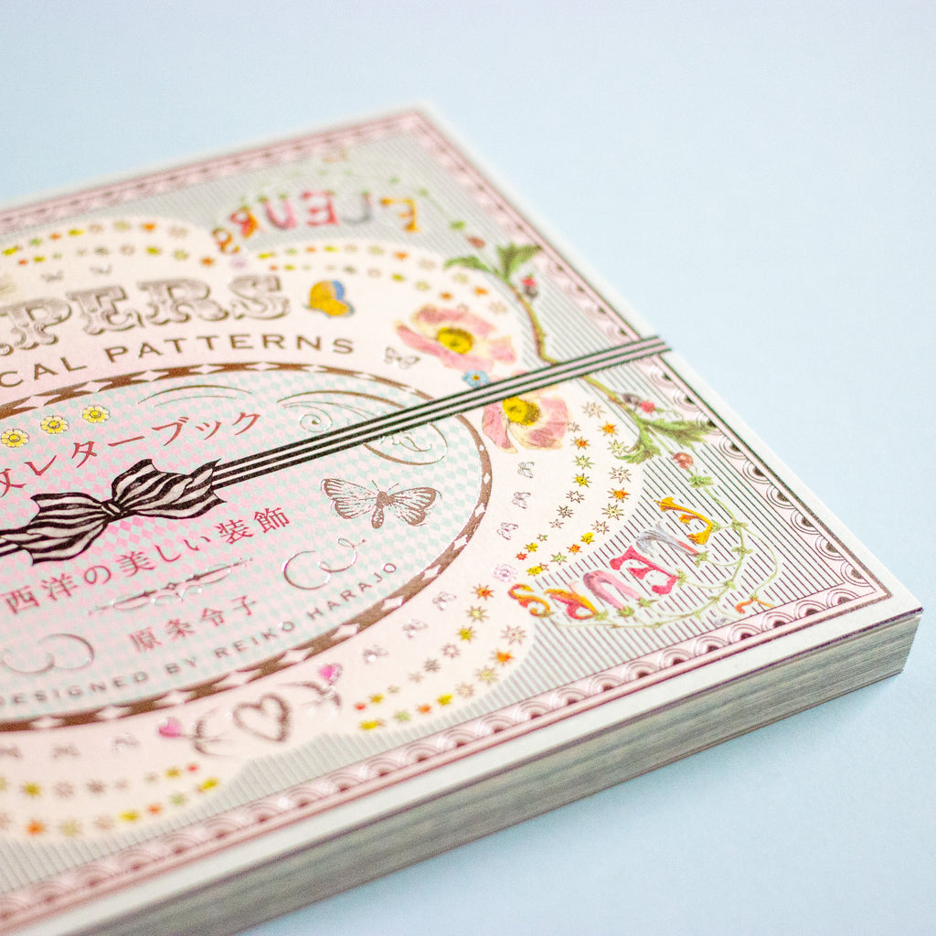 A close-up of a decorative book with Japanese and English text, featuring a pastel floral and ornamental cover design, photographed on a pale blue background.