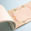An open nbook on a light blue background, showing a beige page with a decorative floral border and colourful letters.