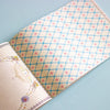 An open book showing a decorative patterned cover with a cream background, blue diamond lattice, and small pink floral motifs, photographed on a light blue surface.