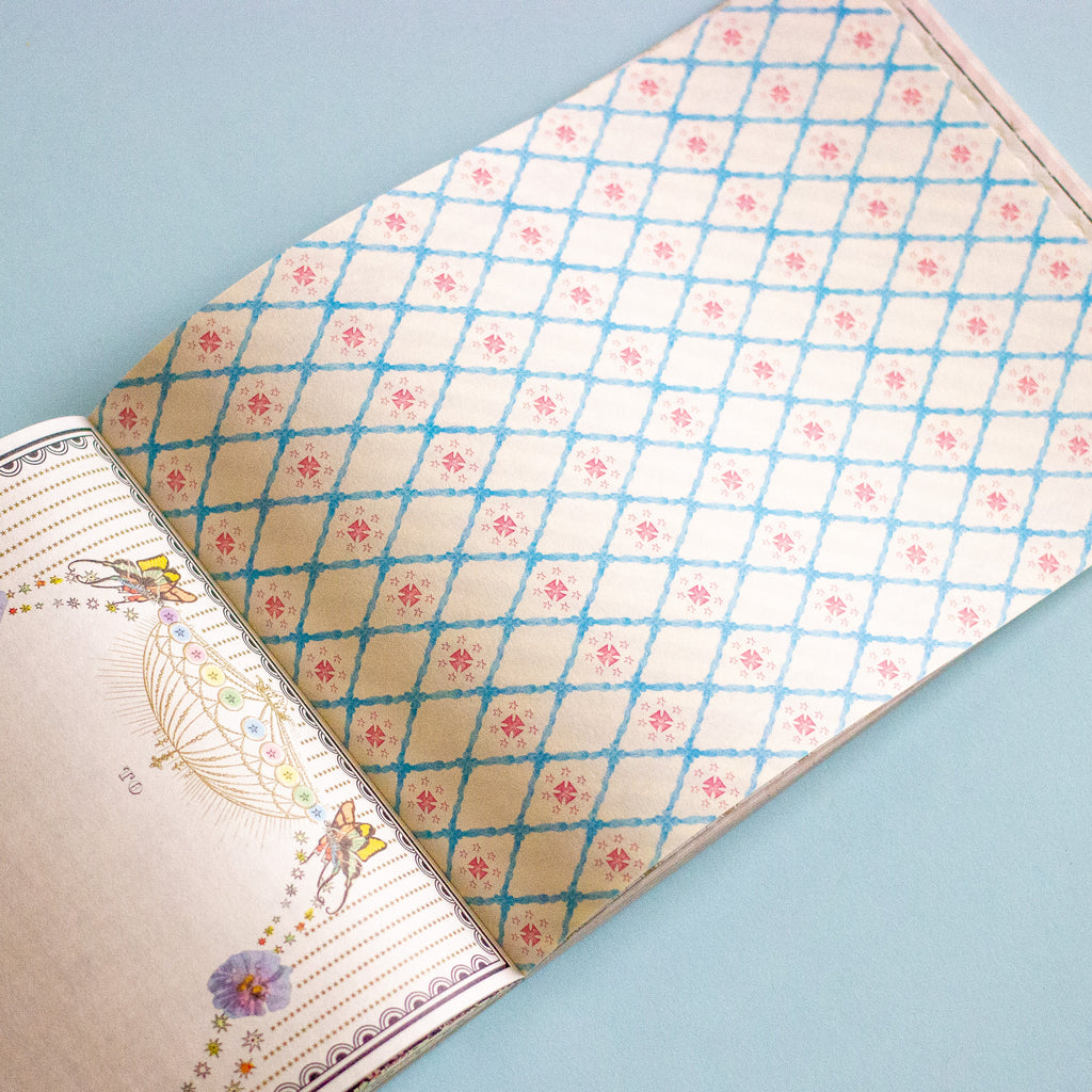 An open book showing a decorative patterned cover with a cream background, blue diamond lattice, and small pink floral motifs, photographed on a light blue surface.