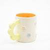 Closeup view of the citrus 3D flower mug handle.  Mug has light glaze with orange interior glaze. Flower shaped handle is citrus coloured. Mug has raised 3D style flowers that are hand painted and glazed in different colours. 