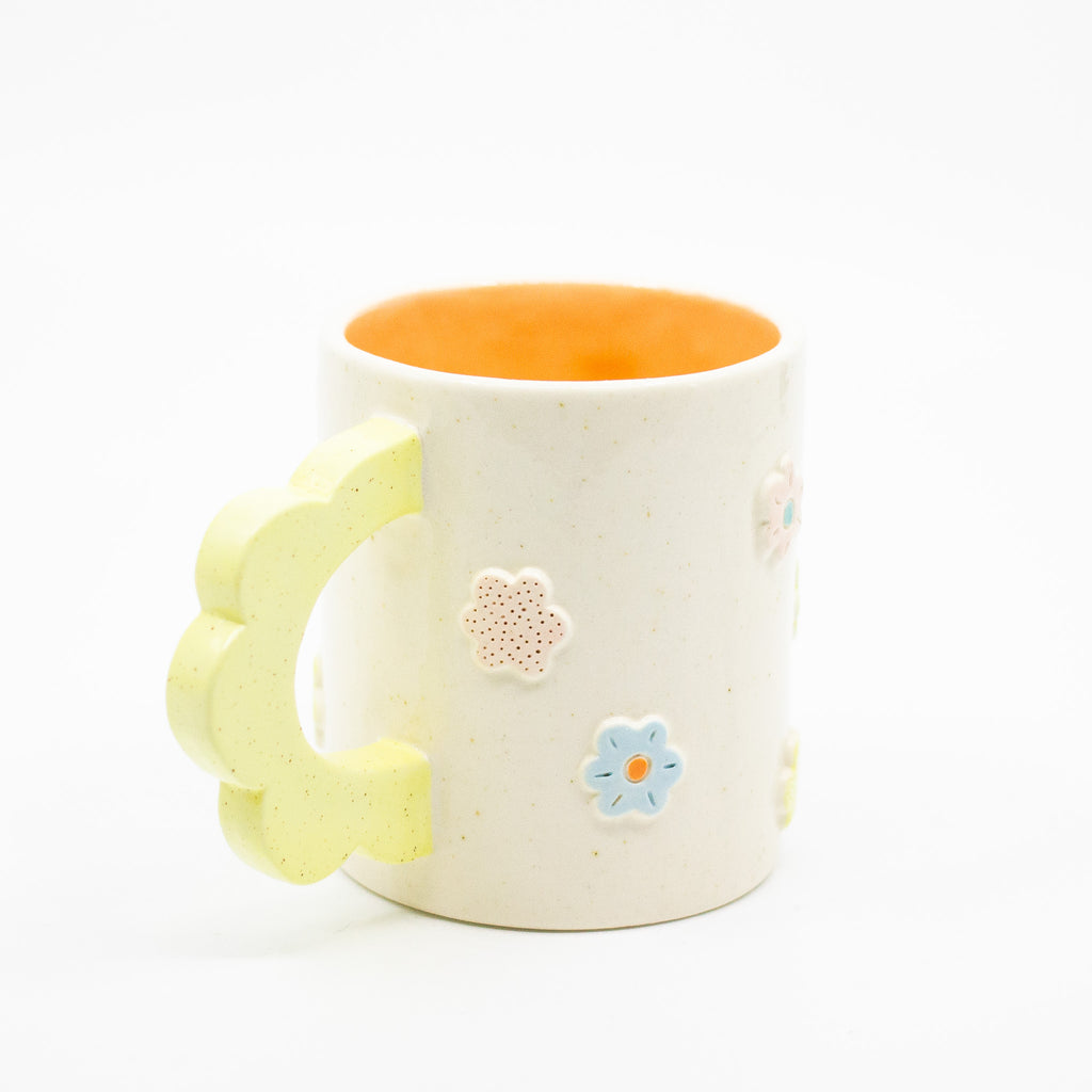 Closeup view of the citrus 3D flower mug handle.  Mug has light glaze with orange interior glaze. Flower shaped handle is citrus coloured. Mug has raised 3D style flowers that are hand painted and glazed in different colours. 