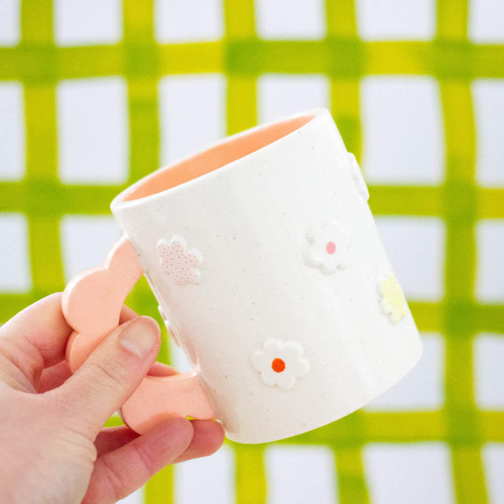 Closeup photo of a ahnd holding the Peach 3D flower mug over a Background of light green and white chequered pattern. Mug has light glaze with orange interior glaze. Flower shaped handle is peach coloured. Mug has raised 3D style flowers that are hand painted and glazed in different colours. 
