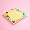 A square yellow sticky note pad with a colourful abstract border design, photographed at an angle on a solid pink background.