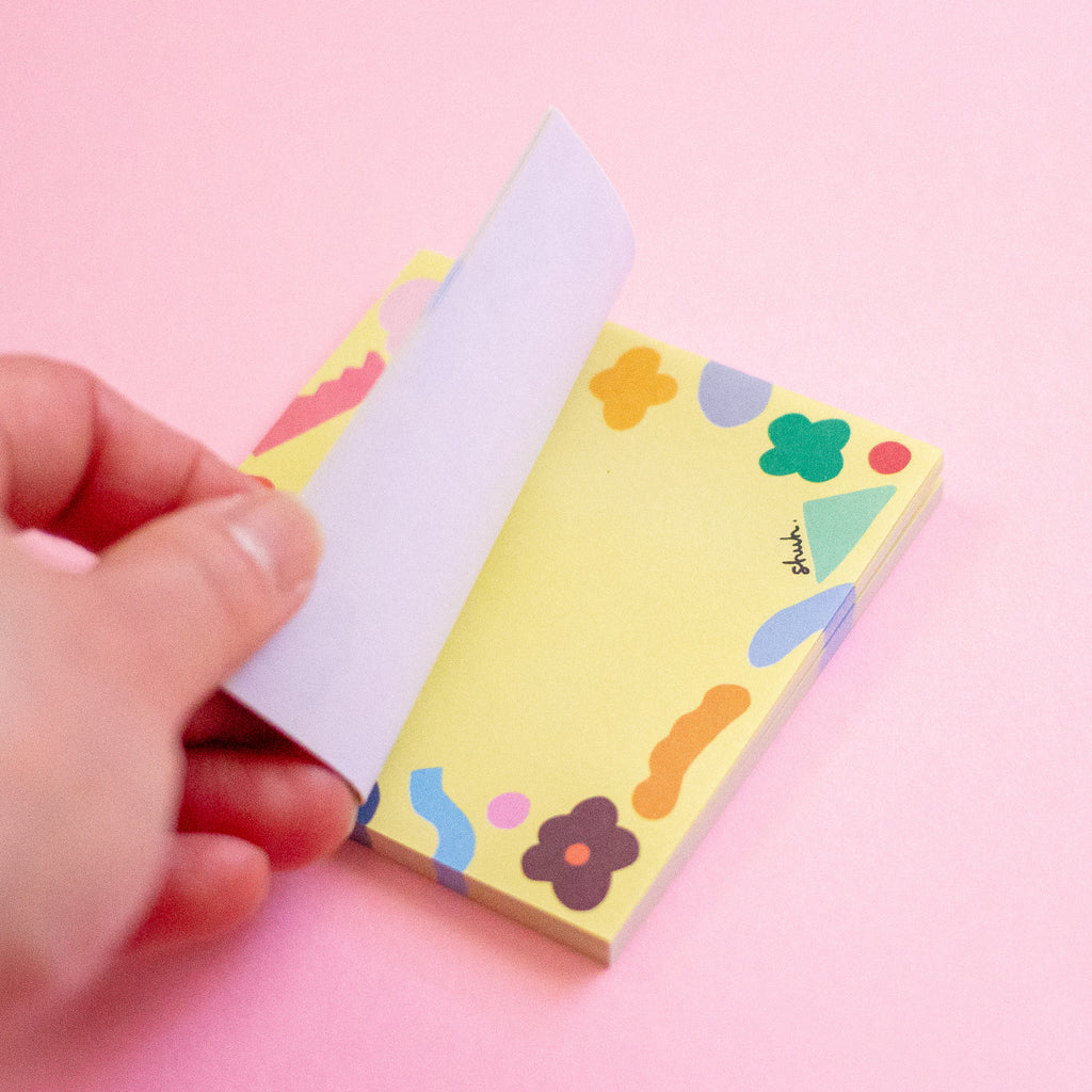 A hand peeling back a page from a yellow sticky note pad decorated with colourful abstract shapes and flowers, set against a solid pink background.