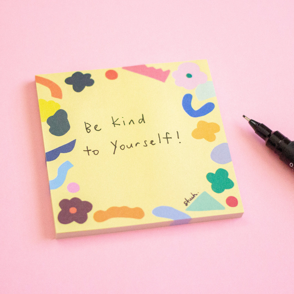 A yellow sticky note pad with the handwritten message 'Be Kind to Yourself!' and colourful abstract shapes around the border, shown on a pink background with a black pen nearby.