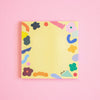 A square yellow notepad with a colourful abstract border of flowers, shapes, and curved lines, photographed flat against a solid pink background. The brand name 'shuh.' is printed in black script near the bottom right corner of the pad.