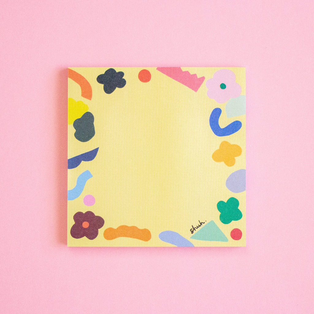 A square yellow notepad with a colourful abstract border of flowers, shapes, and curved lines, photographed flat against a solid pink background. The brand name 'shuh.' is printed in black script near the bottom right corner of the pad.