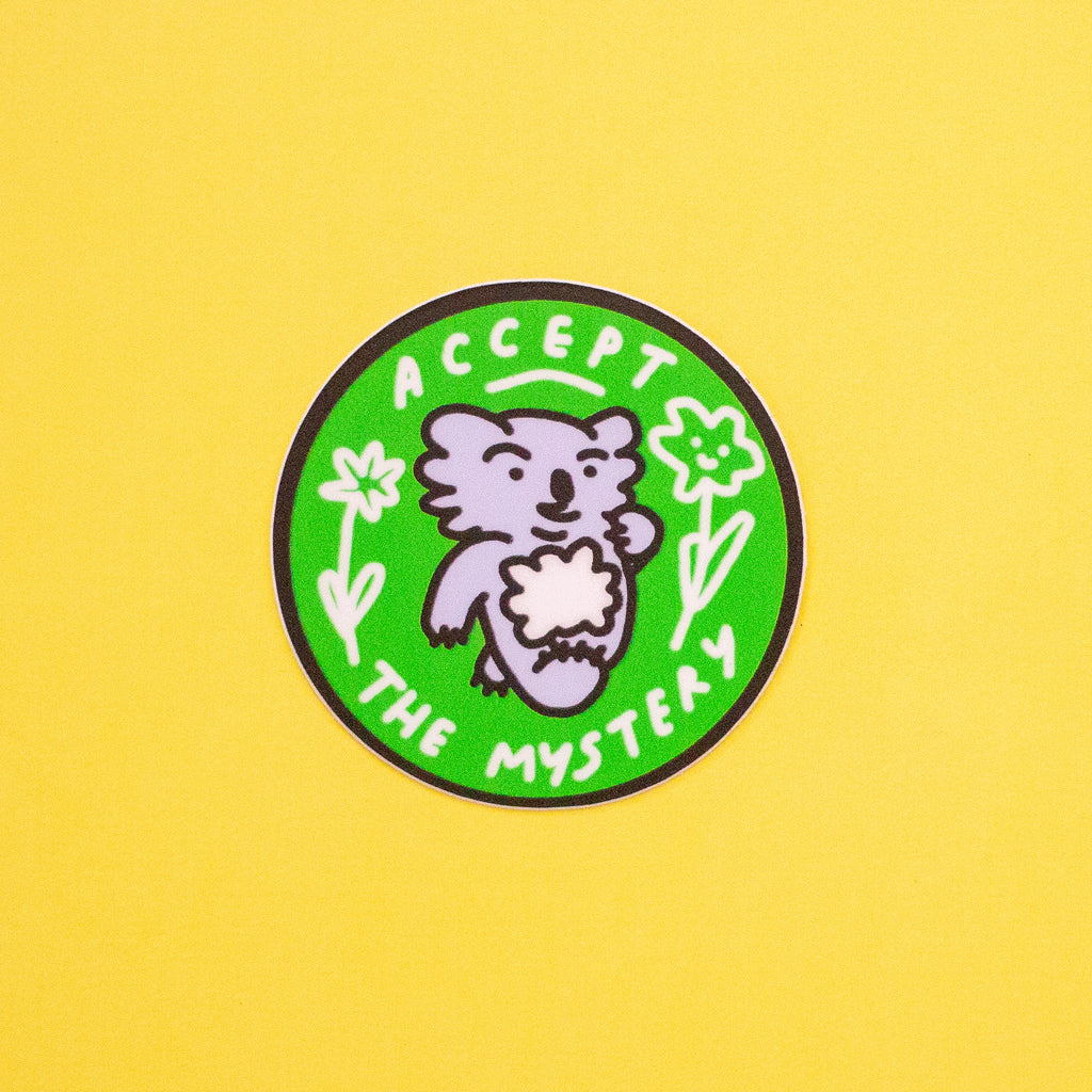 A circular enamel pin featuring a cute koala, set against a bright green background with the text 'ACCEPT THE MYSTERY' in white. The pin is displayed on a solid yellow background.