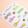 A sheet of colourful sticker designs from the brand Adornments, featuring cute cartoon motifs like a heart-shaped mirror, a pink cartoon, green clovers, and a yellow ribbon banner that reads 'HELLO LOVELY', displayed on a soft peach background.