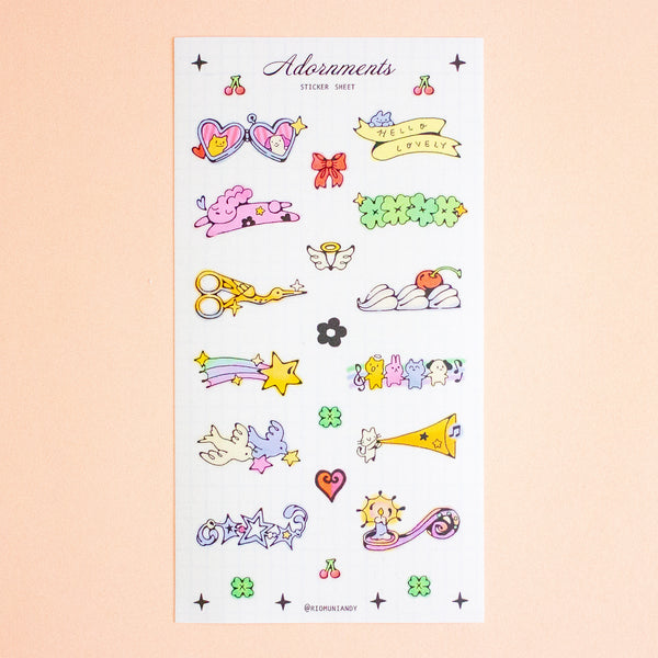 A sticker sheet featuring cute pastel-coluored cartoon illustrations, including cherries, hearts, bows, a green flower, scissors, a rainbow, stars, and small characters, displayed on a soft peach background.