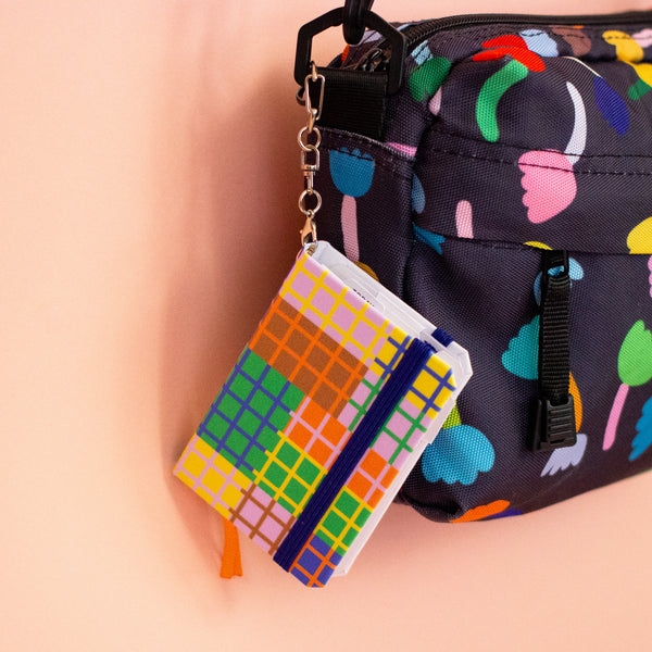 Miniature notebook charm with a colourful grid design, elastic closure, and metal clip keychain. Features an orange ribbon bookmark. Designed by shuh. in collaboration with Ana Tomy, photographed on a peach background.