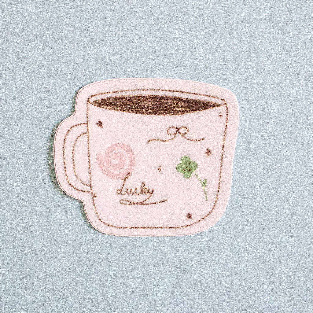 A die-cut sticker shaped like a coffee mug with black outlines, featuring a pink swirl, a green four-leaf clover, and the word 'lucky' in cursive, shown on a light blue background.