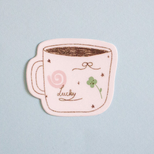 A die-cut sticker shaped like a coffee mug with black outlines, featuring a pink swirl, a green four-leaf clover, and the word 'lucky' in cursive, shown on a light blue background.