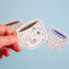 A hand holds a die-cut sticker shaped like a white cup with a pink swirl, a green clover, and the word 'Lucky' in script. Two more blurred stickers appear in the background against a light blue backdrop.
