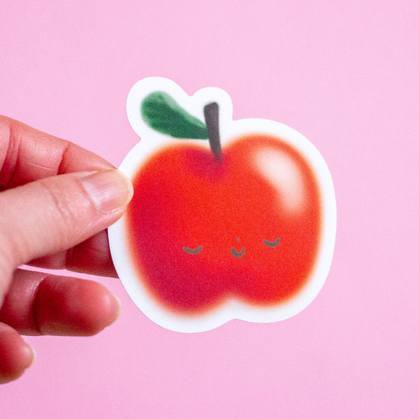 A hand holds a die-cut sticker shaped like a cute red apple with a small green leaf and closed smiling eyes, set against a solid pink background.